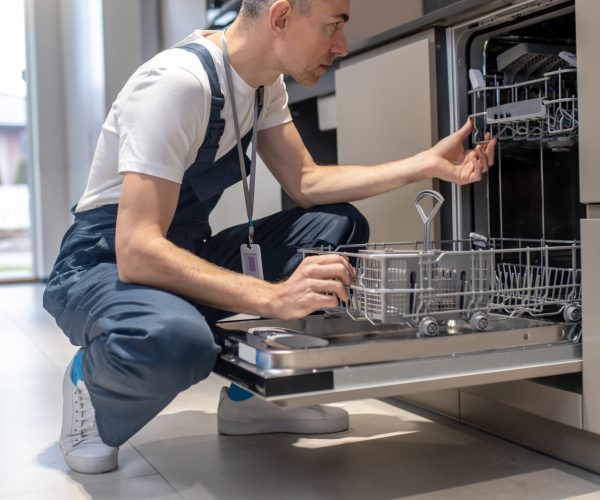 Diagnostics of equipment. Profile of serious man in dark overalls with badge crouching attentively looking at open dishwasher in kitchen at home during day