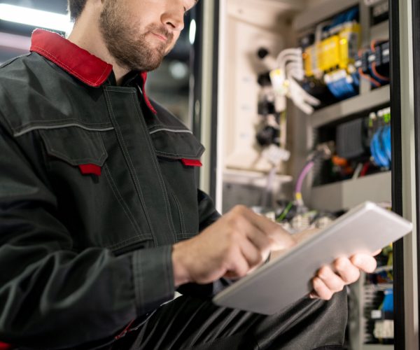 Young bearded engineer in workwear and hardhat checking settings of industrial machine while scrolling through technical data in tablet