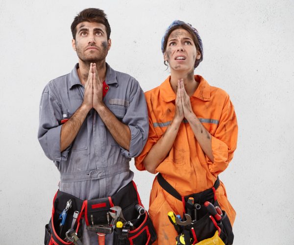 Young female and male plumbers keep hand as pray for rest or holidays, being exhausted after hard work, isolated white background. Dirty maintenance worker keep palms together, look hopefully